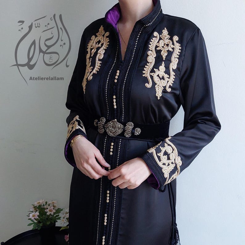Puede incluir: Un vestido de sat&eacute;n negro con bordados dorados y un cintur&oacute;n de terciopelo negro. El vestido tiene un cuello alto y mangas largas. El bordado es intrincado y presenta dise&ntilde;os florales. El cintur&oacute;n tiene una hebilla grande y adornada.