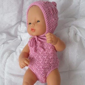 Puede incluir: Una muñeca bebé vestida con un conjunto de punto rosa hecho a mano. El conjunto incluye un gorro, un body y patucos. La muñeca está sobre una superficie blanca. Los artículos de punto son de color rosa liso.
