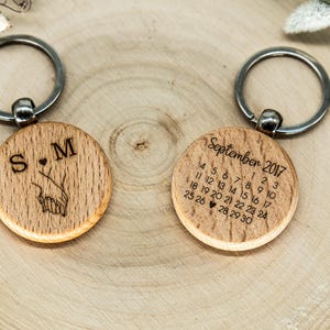 Könnte beinhalten: Zwei runde Holzschlüsselanhänger mit Metallringen. Einer zeigt die eingravierten Buchstaben S und M mit einem kleinen Herz und einer Strichzeichnung von zwei sich haltenden Händen. Der andere zeigt einen Kalender für September 2017 mit einem Herz am 27.