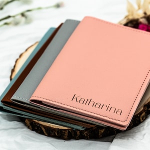 Op de afbeelding: Een stapel gepersonaliseerde paspoorthouders in roze, grijs, bruin en blauwgroen. De roze houder is voorzien van de naam "Katharina". De houders liggen op een houten schijf, tegen een witte stoffen achtergrond.