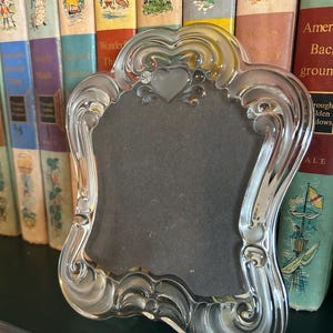 May include: A clear glass picture frame with a decorative, scalloped edge and a heart-shaped detail at the top center. The frame features a smooth, reflective surface and a dark, solid-colored insert within the central display area.