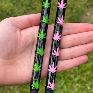 May include: Two black pens with silver tips and black rubber tips. One pen features green cannabis leaf designs, the other pink. Both pens have a glittery finish and are held in a hand against a green background.