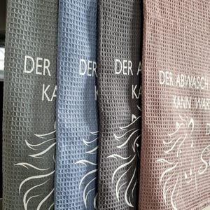 May include: Four kitchen towels with a waffle weave texture. The towels are gray, blue, gray, and pink. Each towel has a white unicorn design and text that reads "Der Abwasch kann warten".