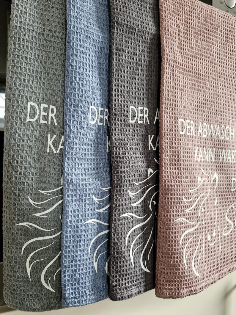 May include: Four kitchen towels with a waffle weave texture. The towels are gray, blue, gray, and pink. Each towel has a white unicorn design and text that reads "Der Abwasch kann warten".