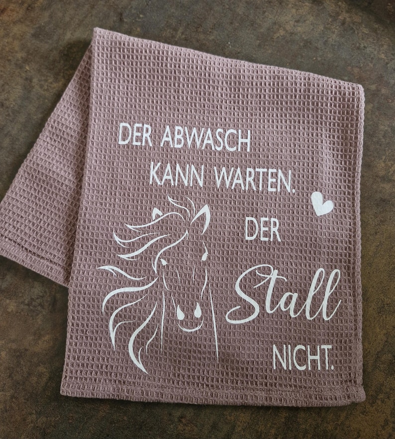 May include: A pink waffle weave kitchen towel with a white horse silhouette and the text "Der Abwasch kann warten. Der Stall nicht."