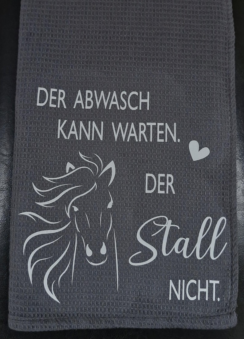 May include: A gray kitchen towel with white text that reads "Der Abwasch kann warten. Der Stall nicht."  A white silhouette of a horse's head is in the center of the towel.