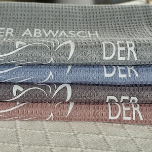 May include: Four waffle weave kitchen towels stacked on top of each other. The towels are gray, blue, gray, and pink. Each towel has the word "DER" printed on it. The top towel has the words "DER ABWASCH" printed on it.