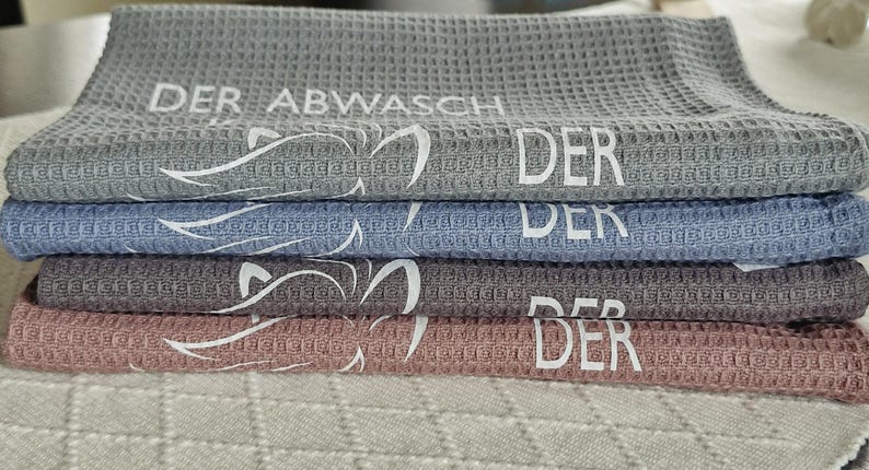 May include: Four waffle weave kitchen towels stacked on top of each other. The towels are gray, blue, gray, and pink. Each towel has the word "DER" printed on it. The top towel has the words "DER ABWASCH" printed on it.