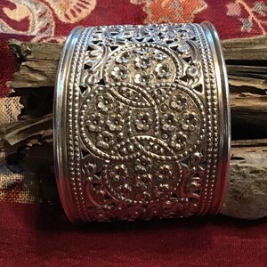 May include: A silver cuff bracelet with intricate floral and geometric patterns. The wide band features detailed cut-out designs and embossed floral motifs. The bracelet is a statement piece, showcasing a vintage or bohemian style.