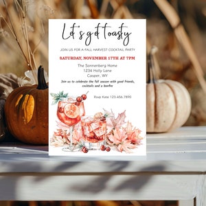Puede incluir: Una tarjeta de invitación con temática otoñal con una ilustración de acuarela de dos cócteles y hojas de otoño. El texto dice "Let's get toasty" e invita a los invitados a una fiesta de cócteles de cosecha de otoño el sábado 17 de noviembre a las 7:00 p. m. en The Sonnenberg Home, 1234 Holly Lane, Casper, WY. La información de RSVP es "Rsvp Kate 123.456.7890".