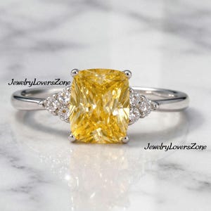 Pode incluir: Um anel de prata com uma grande pedra amarela retangular. Pequenas pedras claras ladeiam a pedra principal. O anel &eacute; exibido sobre um fundo de m&aacute;rmore branco e cinza. O texto "JewelryLoversZone" &eacute; vis&iacute;vel.