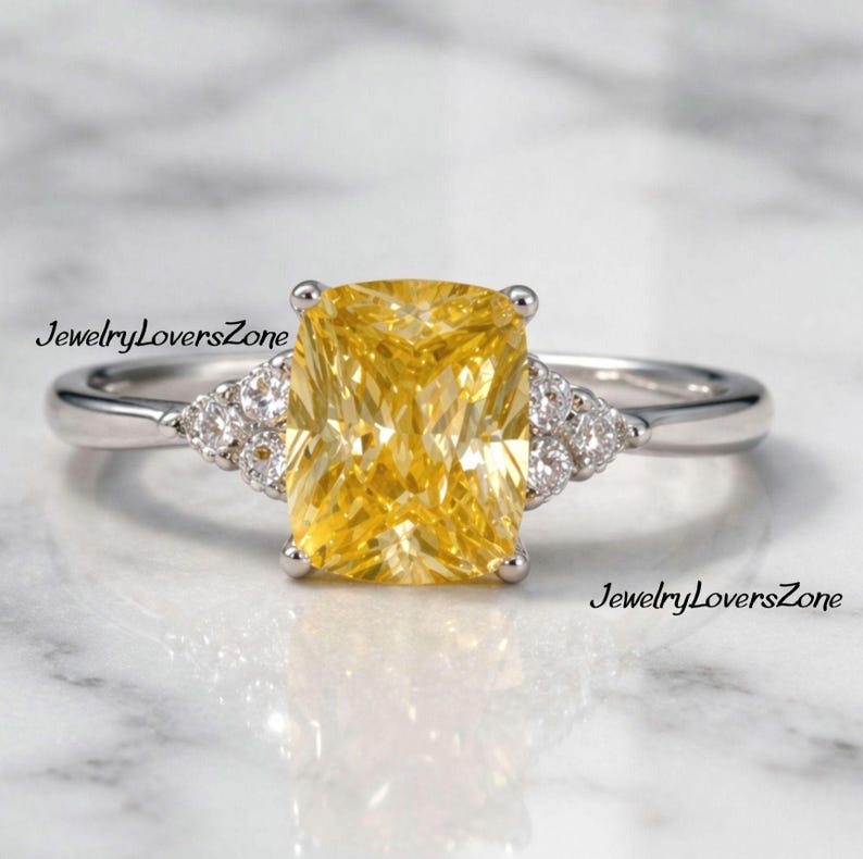 Pode incluir: Um anel de prata com uma grande pedra amarela retangular. Pequenas pedras claras ladeiam a pedra principal. O anel &eacute; exibido sobre um fundo de m&aacute;rmore branco e cinza. O texto "JewelryLoversZone" &eacute; vis&iacute;vel.