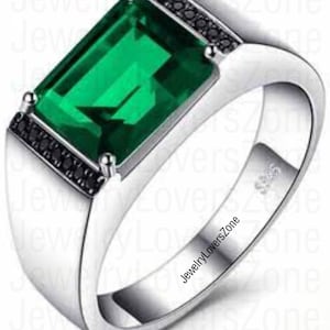 May include: A silver ring with a large, emerald green gemstone set in a rectangular bezel. The ring is accented with small black gemstones around the bezel. The ring is engraved with the text "JewelryLoversZone".