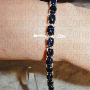 Puede incluir: Un brazalete de plata con una fila de piedras ovaladas de color azul oscuro. Las piedras tienen un patrón de estrella blanca en el centro. El brazalete está en la muñeca de una persona.