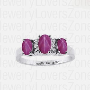Può includere: Un anello d'argento con tre pietre ovali rosa e pietre più piccole trasparenti sulla fascia. L'anello è su uno sfondo bianco. 'JewelryLoversZone' è stampato sull'anello.