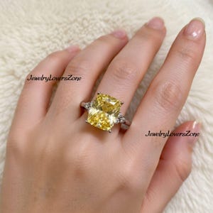Pode incluir: Anel de prata com uma grande pedra amarela retangular. O anel tem pedras menores e transparentes em ambos os lados da pedra principal. O texto "JewelryLoversZone" &eacute; vis&iacute;vel.