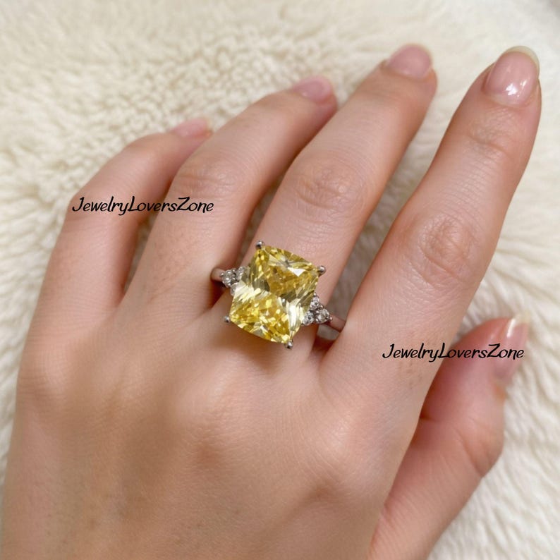 Pode incluir: Anel de prata com uma grande pedra amarela retangular. O anel tem pedras menores e transparentes em ambos os lados da pedra principal. O texto "JewelryLoversZone" &eacute; vis&iacute;vel.