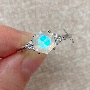 May include: A silver-toned ring featuring a central hexagonal moonstone with a blue iridescent glow, flanked by three small clear stones on each side. The band is thin and metallic, and the text JewelryLoversZone is visible on the side.