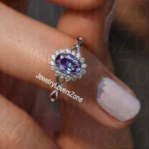 May include: A silver ring featuring a large oval-shaped gemstone set within a halo of smaller diamonds. The gemstone is a deep purple colour with a hint of blue.