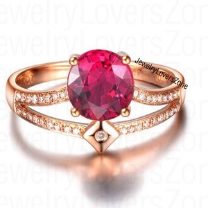 May include: A rose gold ring with a large, round, red gemstone surrounded by smaller, clear gemstones. The ring has a delicate, split band design.