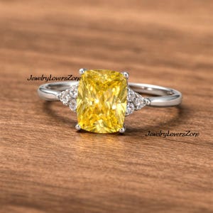 Pode incluir: Um anel de prata com uma grande gema amarela retangular. Pequenas pedras claras adornam os lados da pedra principal. O anel &eacute; exibido sobre um fundo de madeira. O texto "JewelryLoversZone" &eacute; vis&iacute;vel.