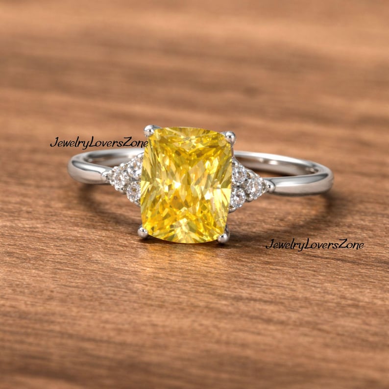 Pode incluir: Um anel de prata com uma grande gema amarela retangular. Pequenas pedras claras adornam os lados da pedra principal. O anel &eacute; exibido sobre um fundo de madeira. O texto "JewelryLoversZone" &eacute; vis&iacute;vel.