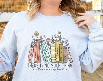 Può includere: Una felpa grigia chiaro con una grafica di una pila di libri colorati con fiori che crescono dalla parte superiore. Il testo "THERE IS NO SUCH THING as too many books" è stampato sotto i libri.