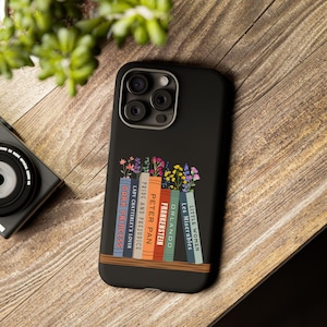 Może przedstawiać: Czarne etui na telefon z wzorem biblioteczki z kwiatami i tytułami klasycznych książek: "Małe kobietki", "Nędznicy", "Orlando", "Frankenstein", "Piotruś Pan", "Dumą i uprzedzeniem", "Kochanek Lady Chatterley" i "Ciemna księżniczka".
