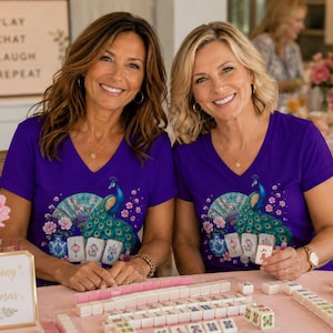 May include: Two purple V-neck t-shirts featuring a graphic print of a blue peacock, a decorative fan, pink flowers, and mahjong tiles. The shirts are worn by two individuals sitting at a table with mahjong tiles and a sign that reads Mahjong & Mimosas.
