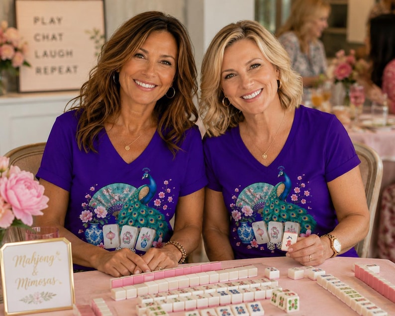 May include: Two purple V-neck t-shirts featuring a graphic print of a blue peacock, a decorative fan, pink flowers, and mahjong tiles. The shirts are worn by two individuals sitting at a table with mahjong tiles and a sign that reads Mahjong & Mimosas.