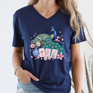 May include: A navy blue V-neck t-shirt featuring a graphic print of a vibrant peacock, a folding fan, three mahjong tiles, two dice, and floral elements including a blue and white vase with pink blossoms and a large pink flower.