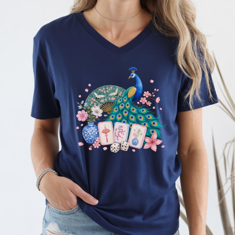 May include: A navy blue V-neck t-shirt featuring a graphic print of a vibrant peacock, a folding fan, three mahjong tiles, two dice, and floral elements including a blue and white vase with pink blossoms and a large pink flower.