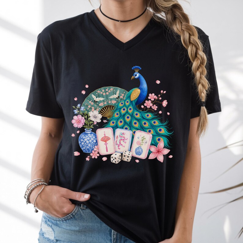 May include: A black V-neck t-shirt featuring a colorful graphic print of a peacock, a folding fan, a blue and white vase, three mahjong tiles, two dice, and pink cherry blossoms. The design is centered on the chest of the short-sleeved garment.