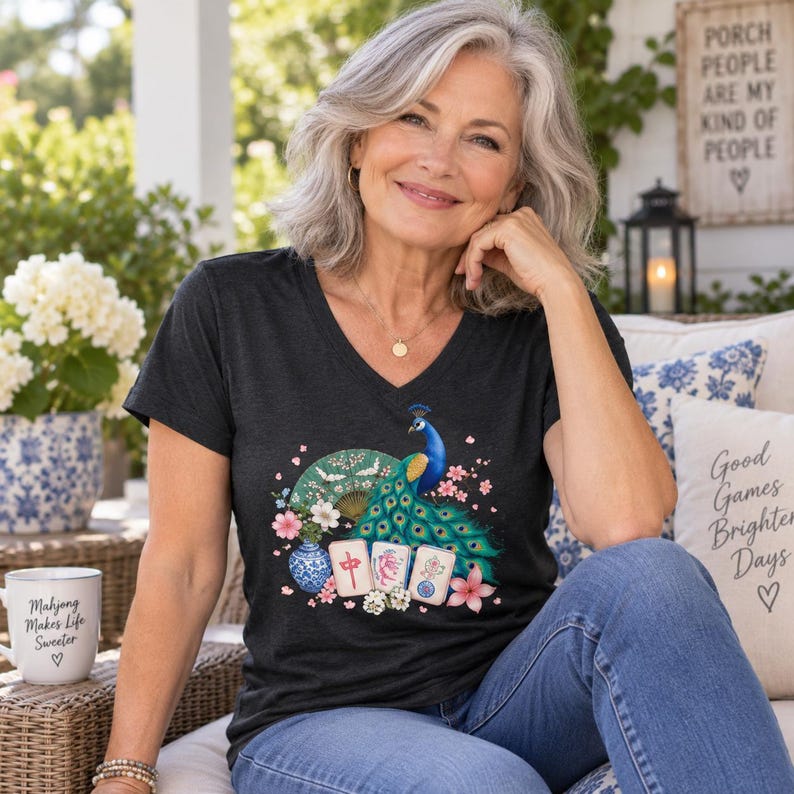 May include: A charcoal gray V-neck t-shirt featuring a graphic print of a blue peacock, a green folding fan, a blue and white patterned vase, pink flowers, and three mahjong tiles. The shirt is worn with blue denim jeans.