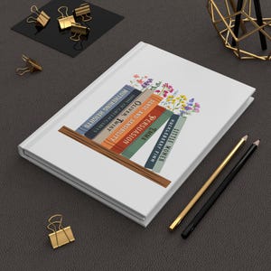 May include: A white journal with a bookshelf design, displaying titles such as "Wuthering Heights" and "Little Women." The design includes colourful flowers above the books. Gold and black pencils and binder clips are also present.