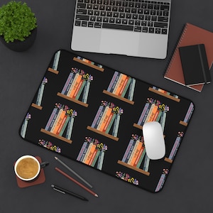 May include: Black mousepad featuring a repeating pattern of bookshelves with colorful books and flowers. The books have titles like "Jane Austen" and "Murdock Mysteries". A white mouse and a laptop are also visible.