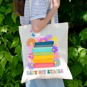 May include: A white canvas tote bag with a colorful illustration of a stack of books surrounded by flowers. The text "BABY GOT STACKS" is printed in rainbow colors at the bottom of the bag.