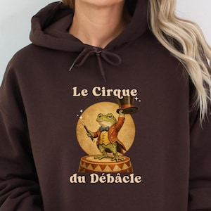 Könnte beinhalten: Dunkelbrauner Kapuzenpullover mit einer Illustration eines Frosches im Zirkus-Outfit. Der Frosch hält einen Zylinder und einen Zauberstab. Der Text lautet "Le Cirque du Débâcle".