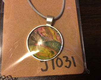 Puede incluir: Un collar de plata con un colgante redondo que presenta remolinos abstractos de pintura rosa, amarilla, verde y dorada. El colgante está encerrado en una resina transparente y tiene un borde plateado. El collar está sobre una tarjeta marrón con el número "J1031" escrito en ella.