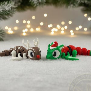 Könnte beinhalten: Zwei skurrile Weihnachtsfiguren. Eine ist ein braunes Rentier mit weißen Geweihen und Flecken. Die andere ist ein grüner Drache mit Weihnachtsmütze und rotem Anzug. Beide stehen vor einem verschneiten Hintergrund mit verschwommenen Lichtern.