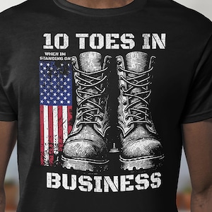 Puede incluir: Camiseta negra con un gráfico de dos botas de trabajo y una bandera estadounidense desgastada. El texto dice "10 TOES IN" encima de las botas y "BUSINESS" debajo, con la frase "WHEN IM STANDING ON" en medio.