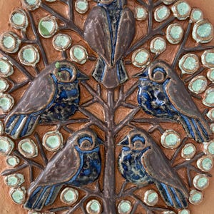May include: A ceramic tile featuring a tree with five birds. The birds have blue and brown coloring, and the tree has brown branches and green circular leaves. The background is a light brown color.