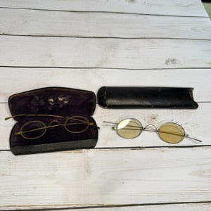 May include: Two pairs of antique eyeglasses with round lenses. One pair is in a black, velvet-lined case, and the other is next to a black leather case. The lenses are a light yellow color, and the frames are a thin, gold-colored metal.