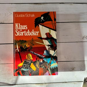 May include: A vintage book titled "Klaus Störtebeker" by Gustav Schalk. The cover features a pirate ship with a skull and crossbones flag, and illustrations of pirates in action. The book has an orange cover with white text.