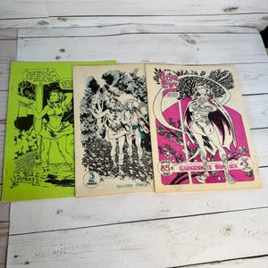 May include: Three vintage comic books are displayed, each with unique cover art. The covers feature illustrations in shades of green, black, white, and pink. The comic books are labeled with text, including the words "2 Big Stories", "Second Issue", and "Glamazon's Burden".