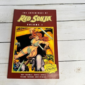 Może przedstawiać: Komiks zatytułowany "The Adventures of Red Sonja Volume I" z żywą ilustracją wojowniczki władającej mieczami. Okładka zawiera tekst "Featuring Conan!" oraz nazwiska autorów.