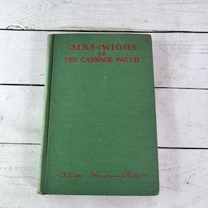 May include: A vintage hardcover book titled "Mrs. Wiggs of the Cabbage Patch" with a green cover and red lettering. The author's name is printed in red at the bottom. The book is set against a white wooden background.