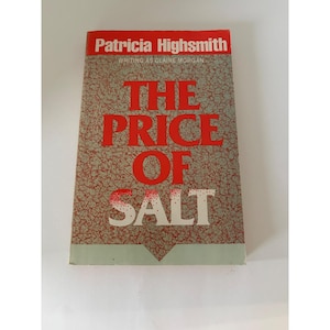 May include: A paperback book titled The Price of Salt by Patricia Highsmith, writing as Claire Morgan. The cover features a textured grey and red marbled background with bold red lettering for the title and white text for the author name.