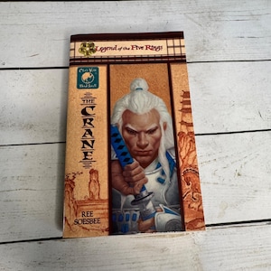 May include: A paperback book titled "The Crane" from the "Legend of the Five Rings" series. The cover features a warrior with white hair and a blue sword. The book has a tan and brown color scheme.