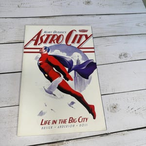 May include: Astro City comic book cover featuring a superhero in a red and blue suit with a flowing cape, soaring above a city skyline. The title is in red and white, with the text "Life in the Big City" below.